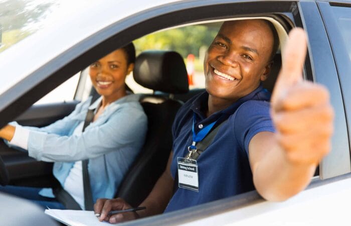 uma mulher dirige o carro, um homem sorri e manda sinal de positivo pela janela